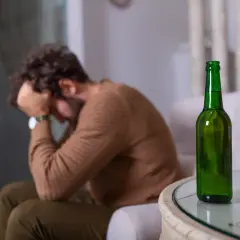 Mężczyzna z głową w dłoniach siedzi obok pustej butelki piwa. Może to symbolizować uczulenie na alkohol lub jego nadużywanie.
